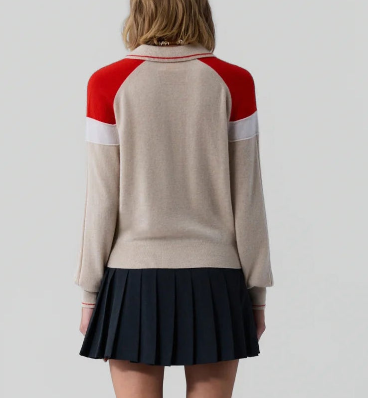 A person stands facing away, wearing the Guest in Residence Alpine Collegiate Polo—a women's cashmere sweater in beige with red and white accents—and a black pleated skirt, set against a plain light gray background.