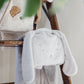 The Bloom Tuck Me in Blanket by Bunnies By the Bay, a plush grey bunny blanket with floppy ears, elegantly drapes over a wooden ladder—ideal as a baby shower gift. Another furry item peeks above, while green leaves add a natural touch in the foreground.