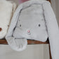 The Bloom Tuck Me in Blanket from Bunnies By the Bay, featuring a gray plush bunny with droopy ears and a pink nose resting on a wooden shelf, is an ideal baby shower gift.