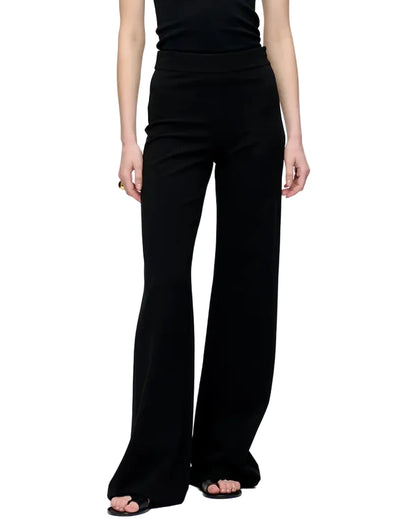 Model wears the Anouk Pant by Callas Milano, featuring a high-waisted, wide-leg crepe jersey design, paired with a sleeveless black top against a plain white background.