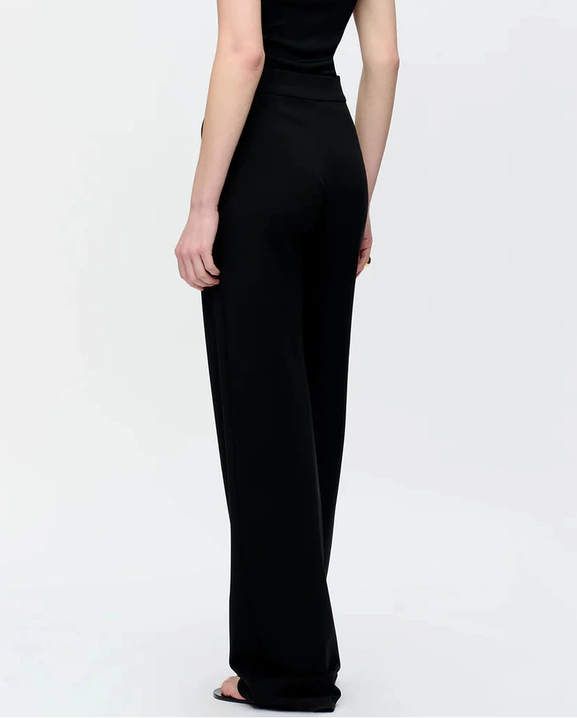 A person stands facing away, wearing Callas Milano's Anouk Pant in black crepe jersey and a sleeveless black top against a plain light background.