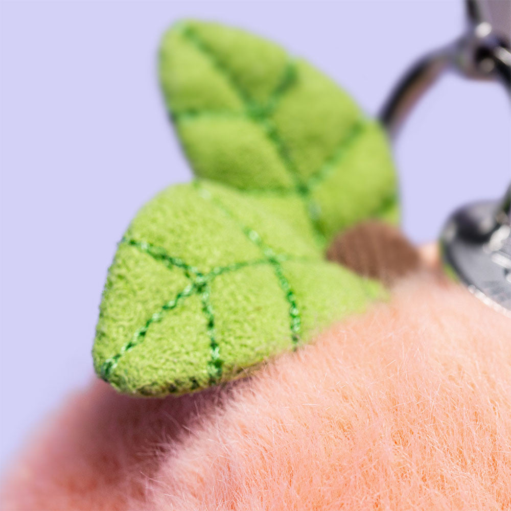 Close-up of the Jellycat Amuseables Peach Bag Charm, showing two green fabric leaves and a metal keyring, set against a light purple background.