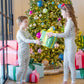 Two kids in matching Lila & Hayes Kids' Grayson Pajama Pant Sets exchange a wrapped gift in front of a decorated Christmas tree with presents underneath.