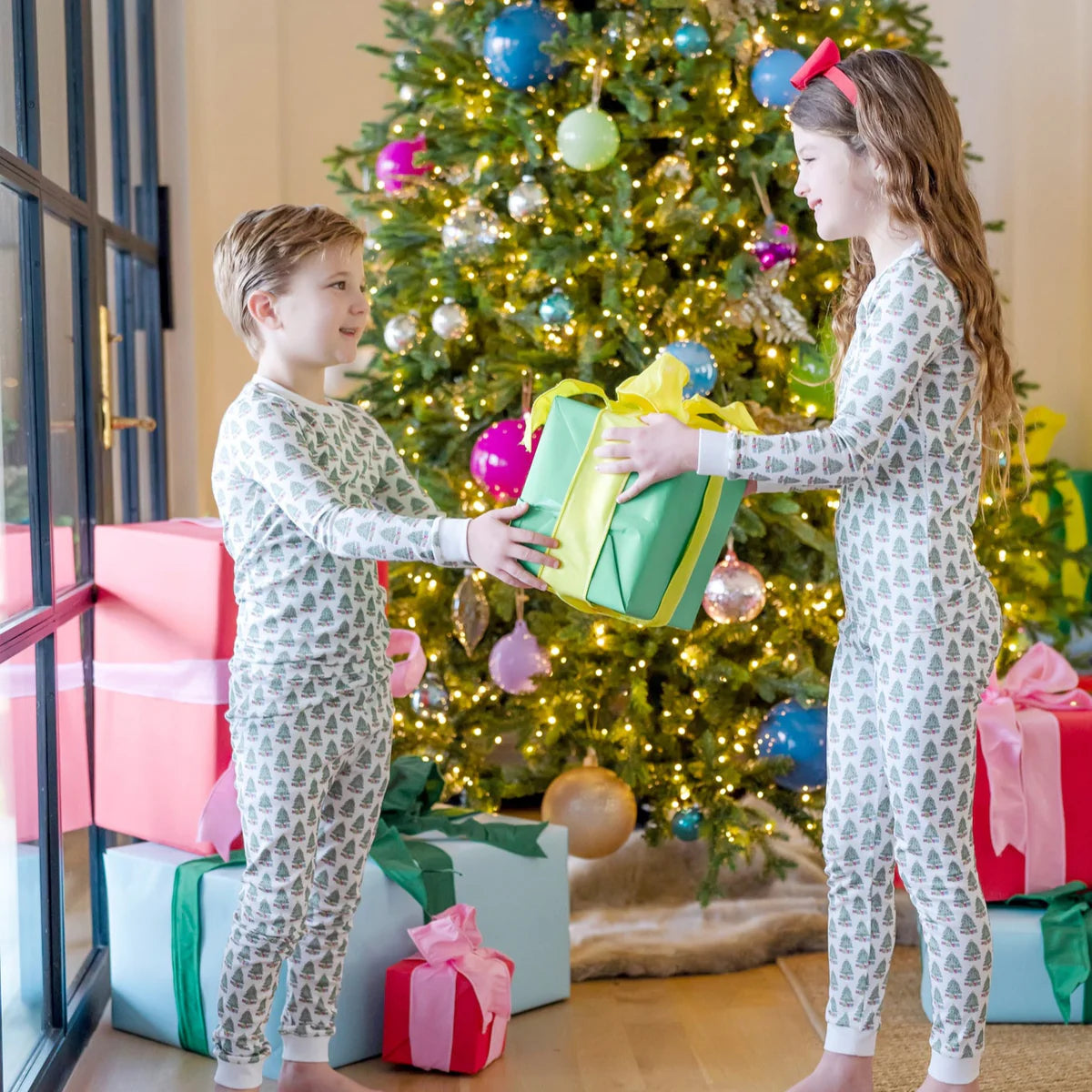 Two kids in matching Lila & Hayes Kids' Grayson Pajama Pant Sets exchange a wrapped gift in front of a decorated Christmas tree with presents underneath.