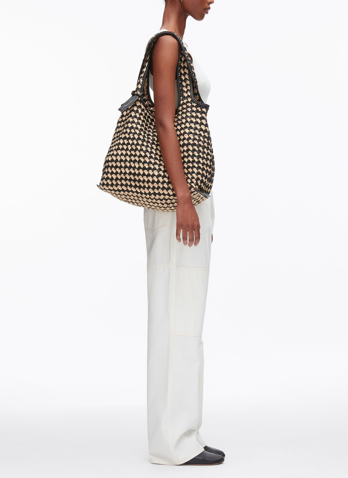 A person in a white sleeveless top and white pants stands in profile, carrying the 3.1 Phillip Lim Crochet Raffia Market Tote over their shoulder.