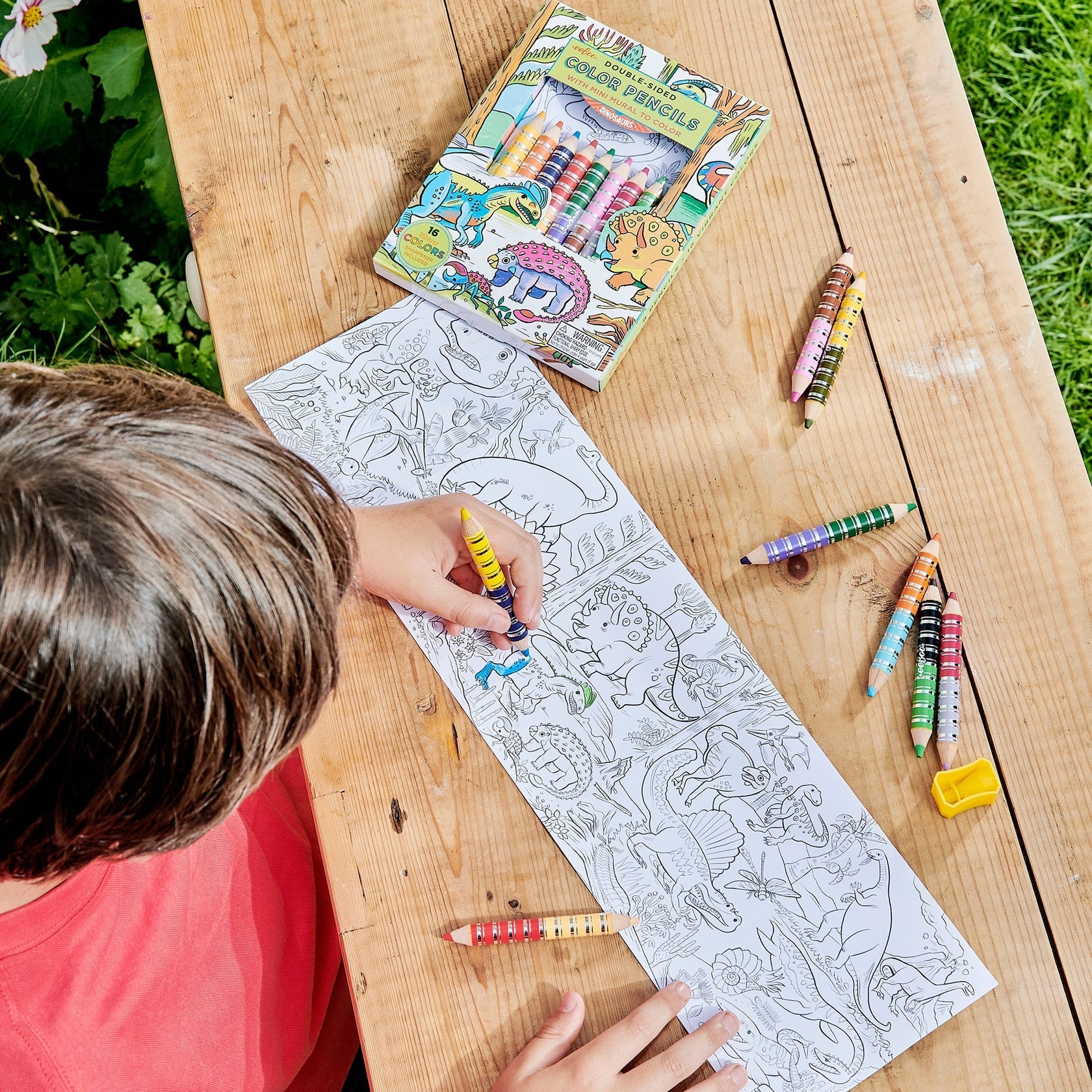 A child colors an Eeboo Mini Mural with Color Pencils at a wooden table; a box of crayons and loose crayons nearby evoke the wonder of magical creatures.
