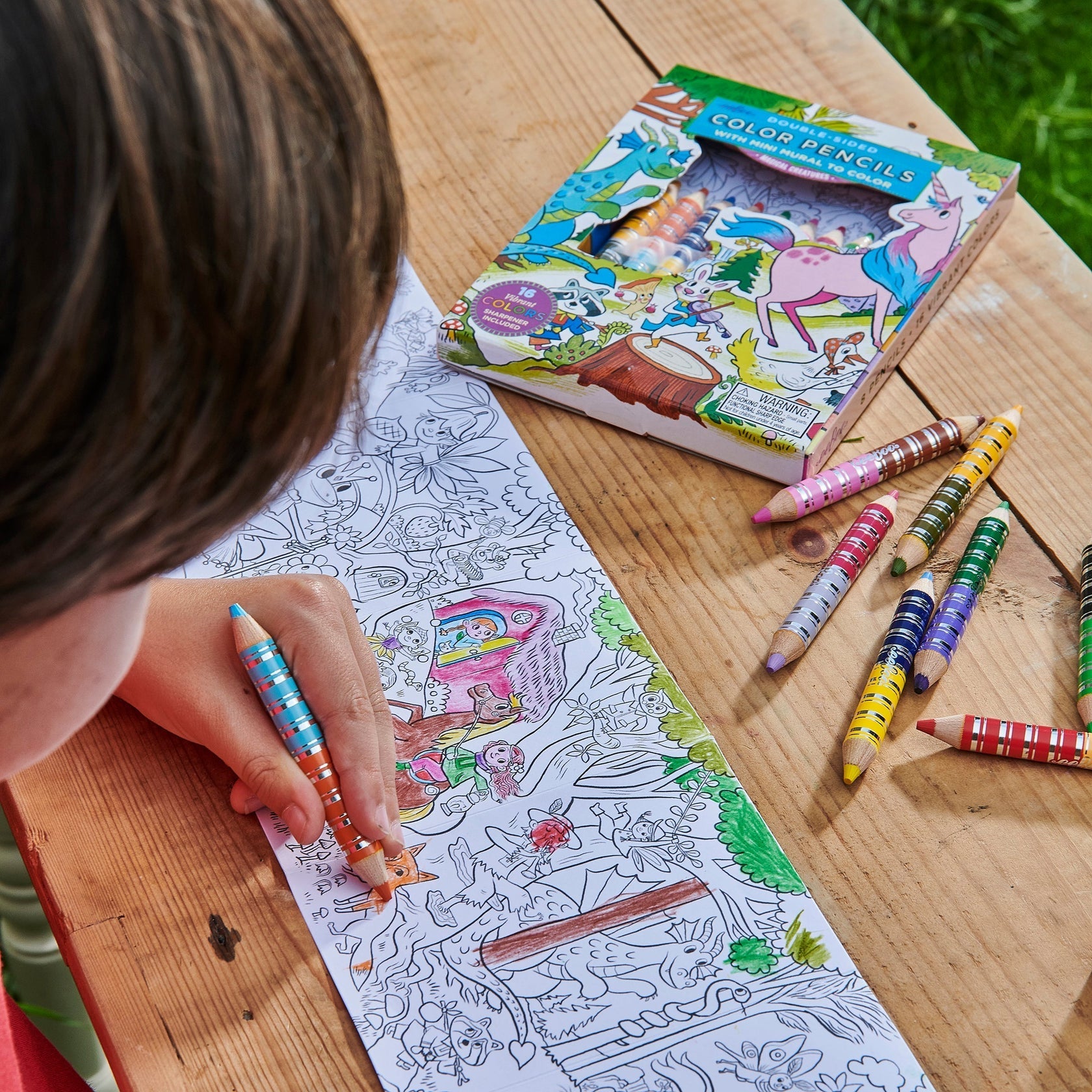 A child colors the Eeboo Mini Mural with Color Pencils, featuring magical creatures, using striped color pencils, while a box of color pencils lies scattered on a wooden table nearby.