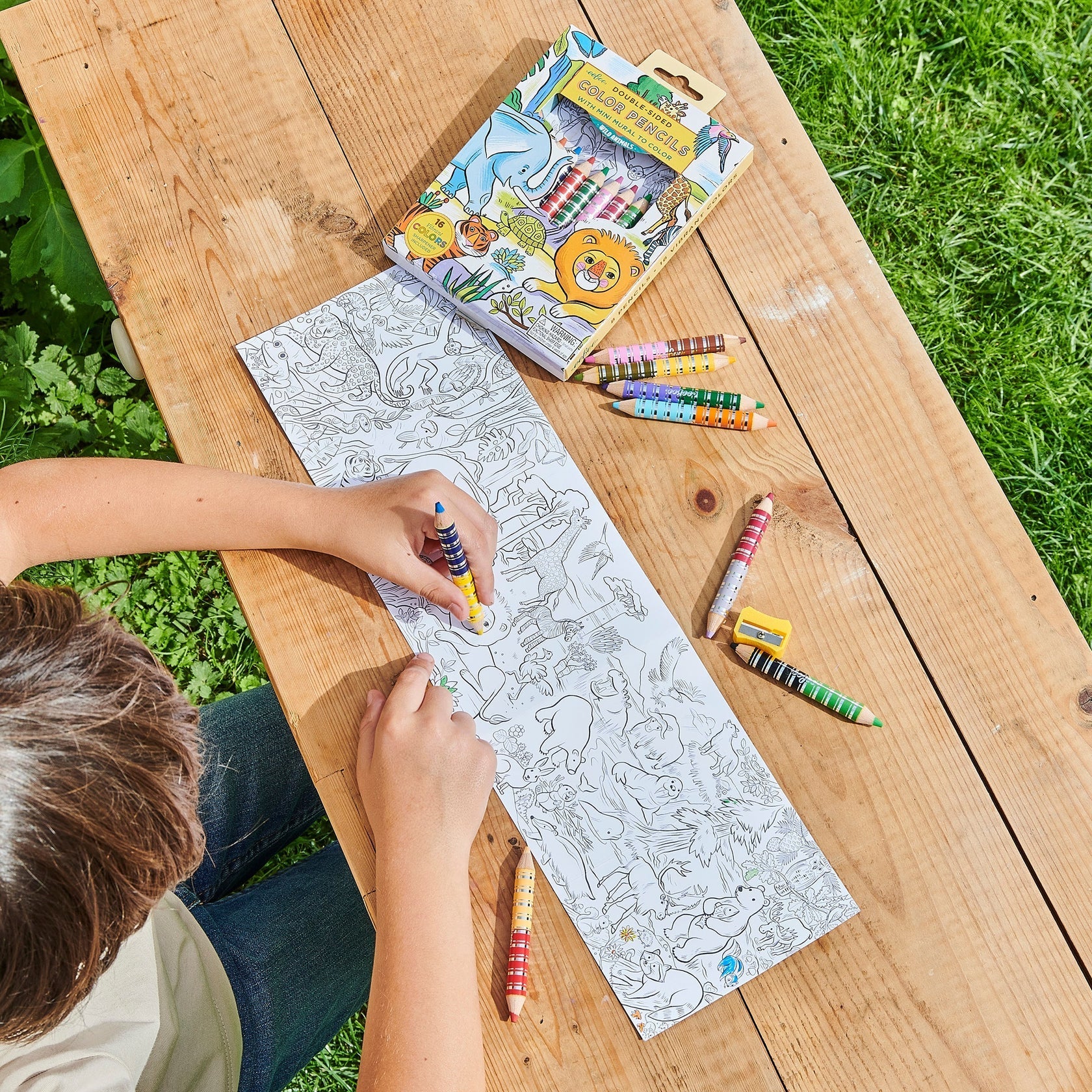 A child colors an animal-themed Eeboo Mini Mural with color pencils at a wooden table outdoors, beside a box of pencils and grass.