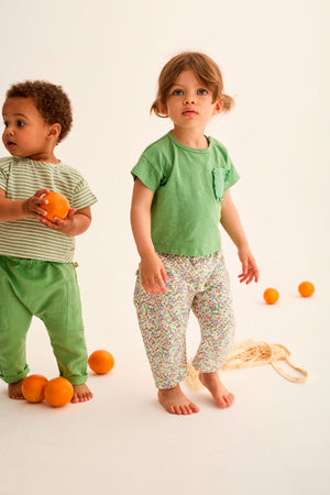 Two young children stand barefoot on a light background with oranges on the floor. One wears green cotton pants and a striped shirt, while the other wears a green top and Cozmo's Baby Girls' Atena Floral Pants.