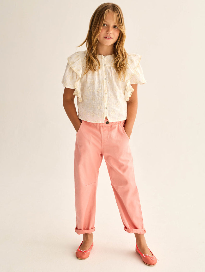 A young girl stands with her hands in her pockets, wearing a white ruffled blouse, Cozmo Girls' Atena Pants featuring a classic twill design and elastic waistband, paired with pink flats against a neutral background.