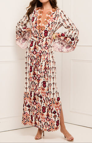 A person stands indoors against a white wall wearing the Figue Athena Dress, a long maxi dress with patterns, bell sleeves, and a tie waist.