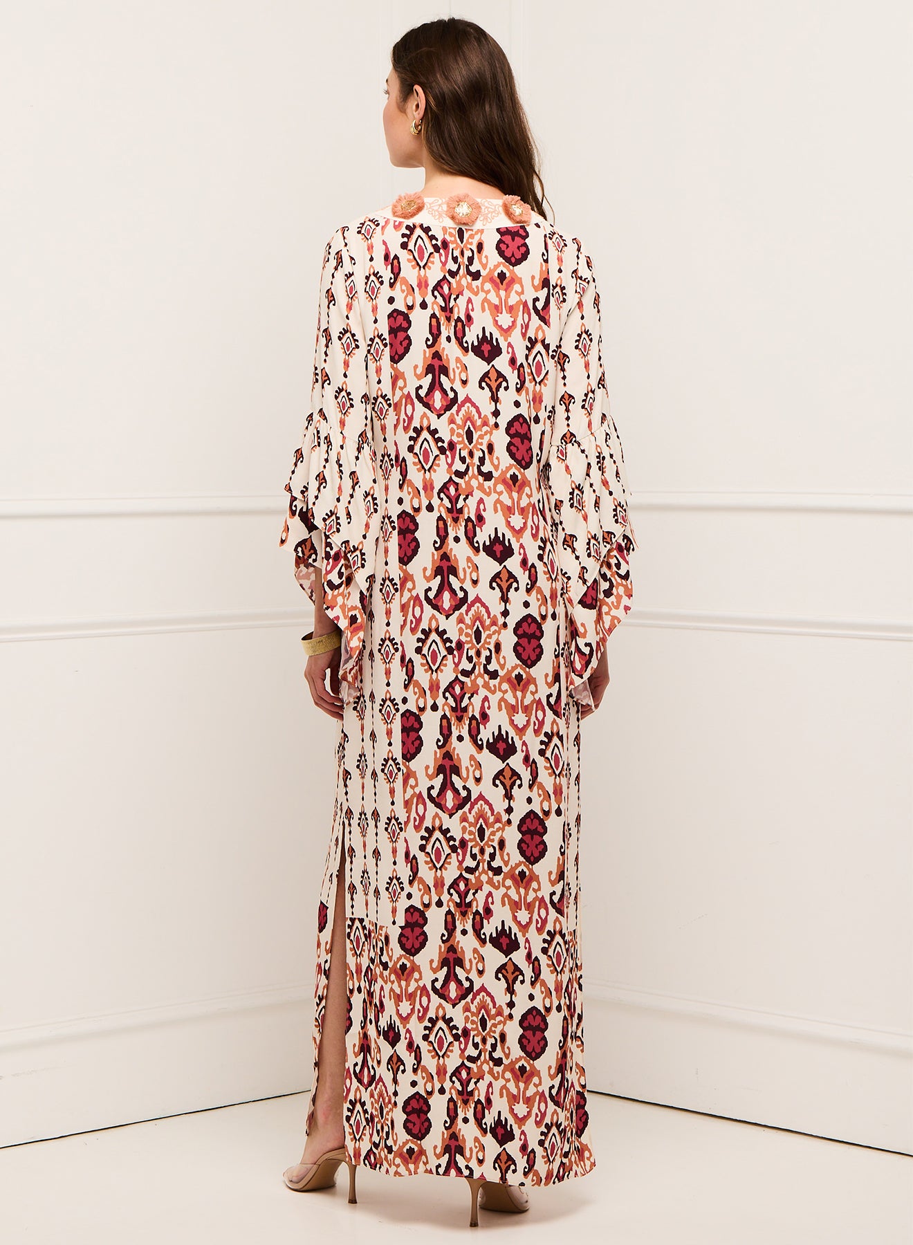 A woman faces away in a light room, wearing the Figue Athena Dress—a long, patterned piece with wide sleeves, side slits, and red, pink, and beige designs.