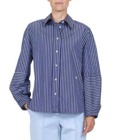 A person is wearing the Aura Top by Callas Milano, a blue and white Italian stripe poplin button-up shirt, paired with light blue pants against a plain white background.