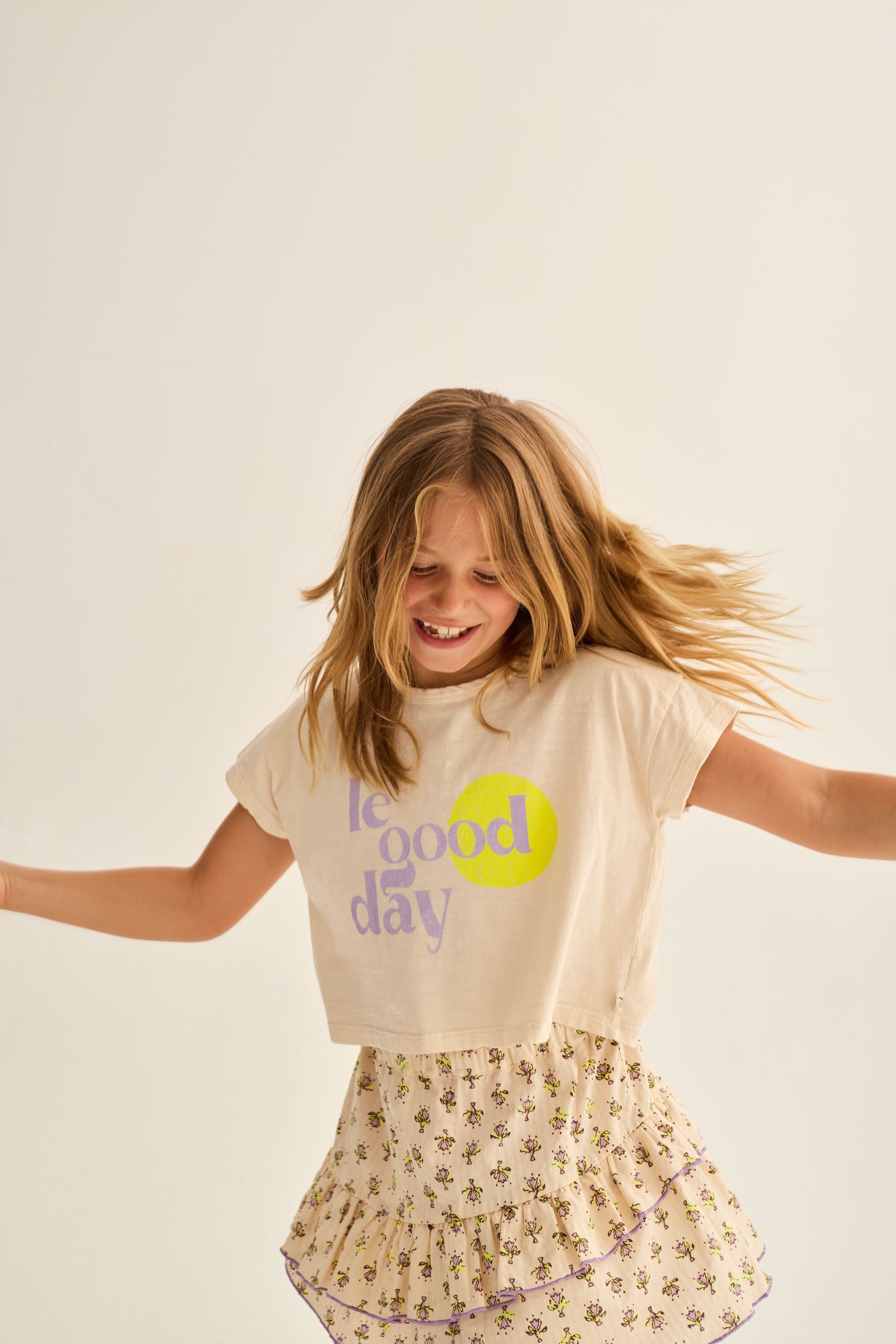 A young girl with long hair, wearing a “feel good day” T-shirt and the Cozmo Girls' Molly Flower Print Skirt, smiles and twirls against a plain light background.