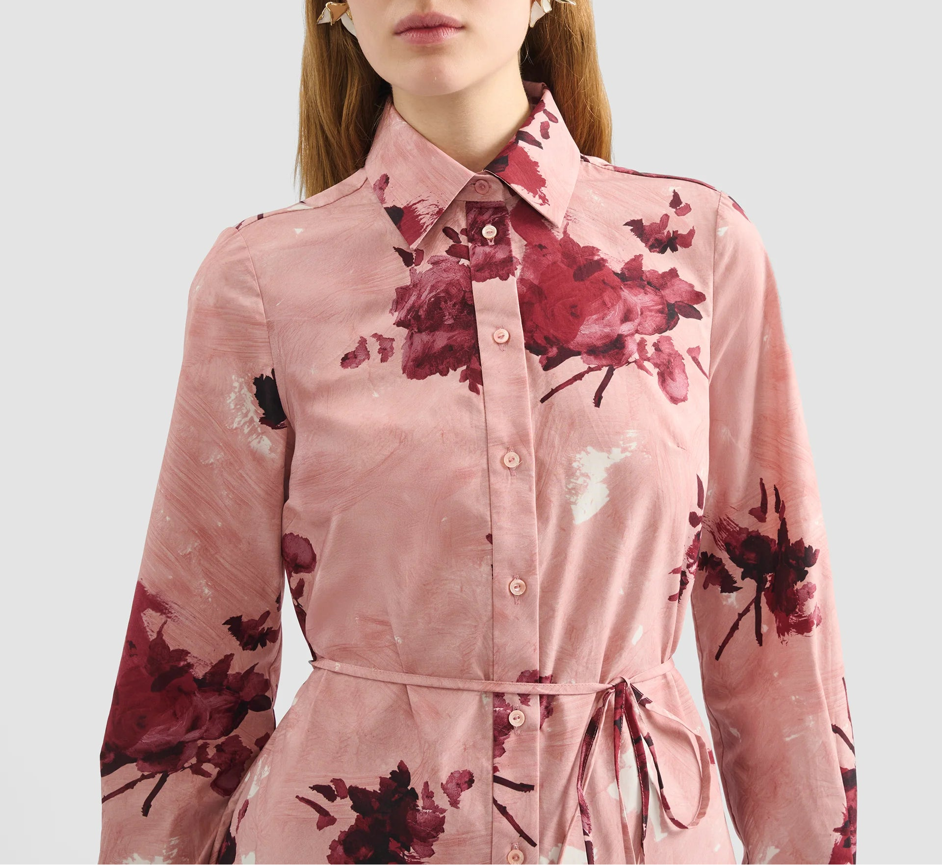 A woman wears the Erdem Long Sleeve Abstract Rose Shirt Dress, a pink cotton poplin button-up with rose bouquets and waist tie, standing against a plain light background.