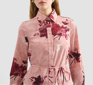 A woman wears the Erdem Long Sleeve Abstract Rose Shirt Dress, a pink cotton poplin button-up with rose bouquets and waist tie, standing against a plain light background.
