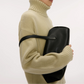 A person in a beige turtleneck holds a large black Mansur Gavriel Helios Bag under their arm, its sleek leather and elegant shoulder straps standing out against a plain background.