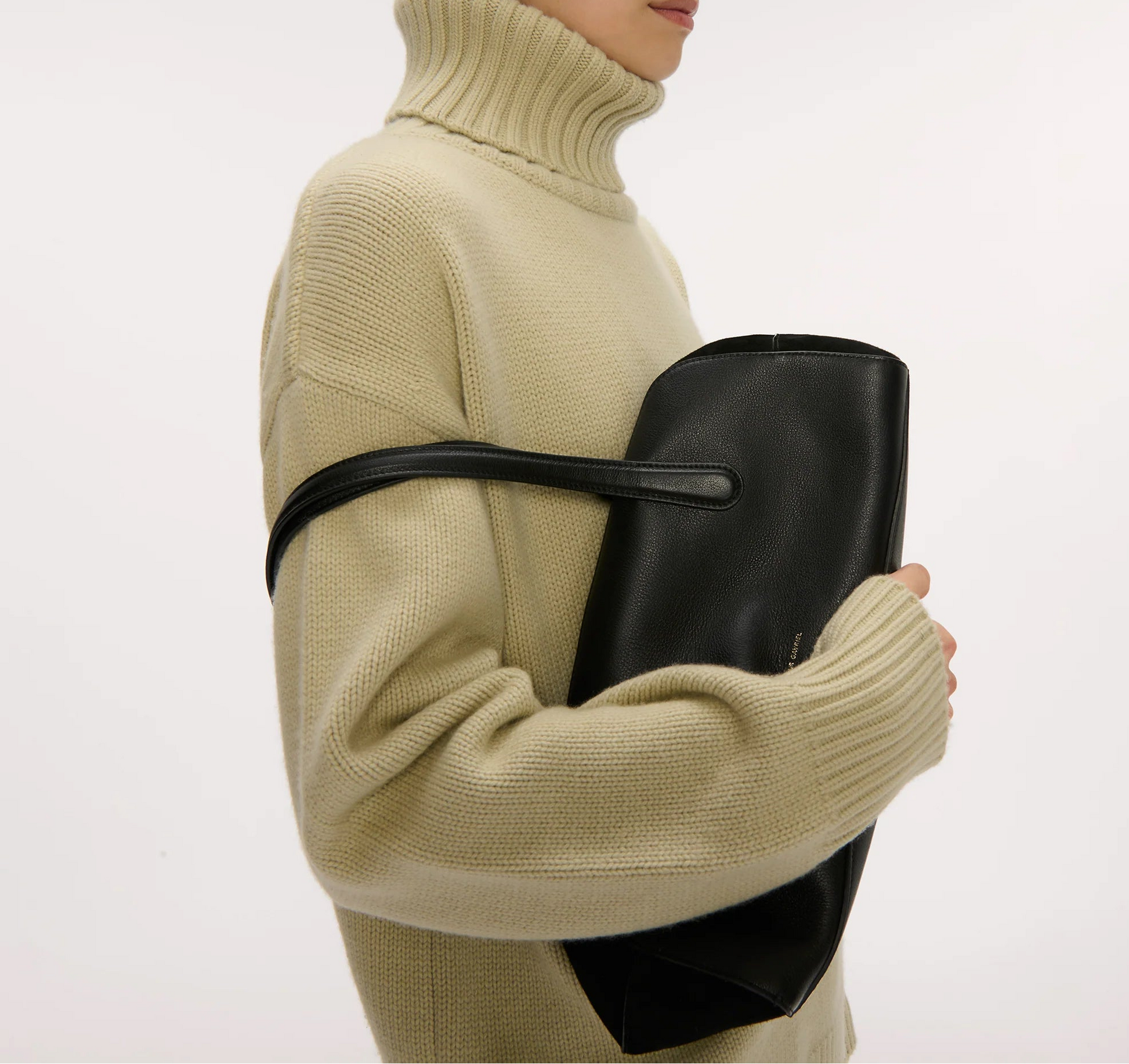 A person in a beige turtleneck holds a large black Mansur Gavriel Helios Bag under their arm, its sleek leather and elegant shoulder straps standing out against a plain background.