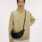 A woman in a beige turtleneck sweater and white pants stands with one hand on her hip, carrying the Mini Fortuna Bag by Mansur Gavriel, crafted from premium calf leather.