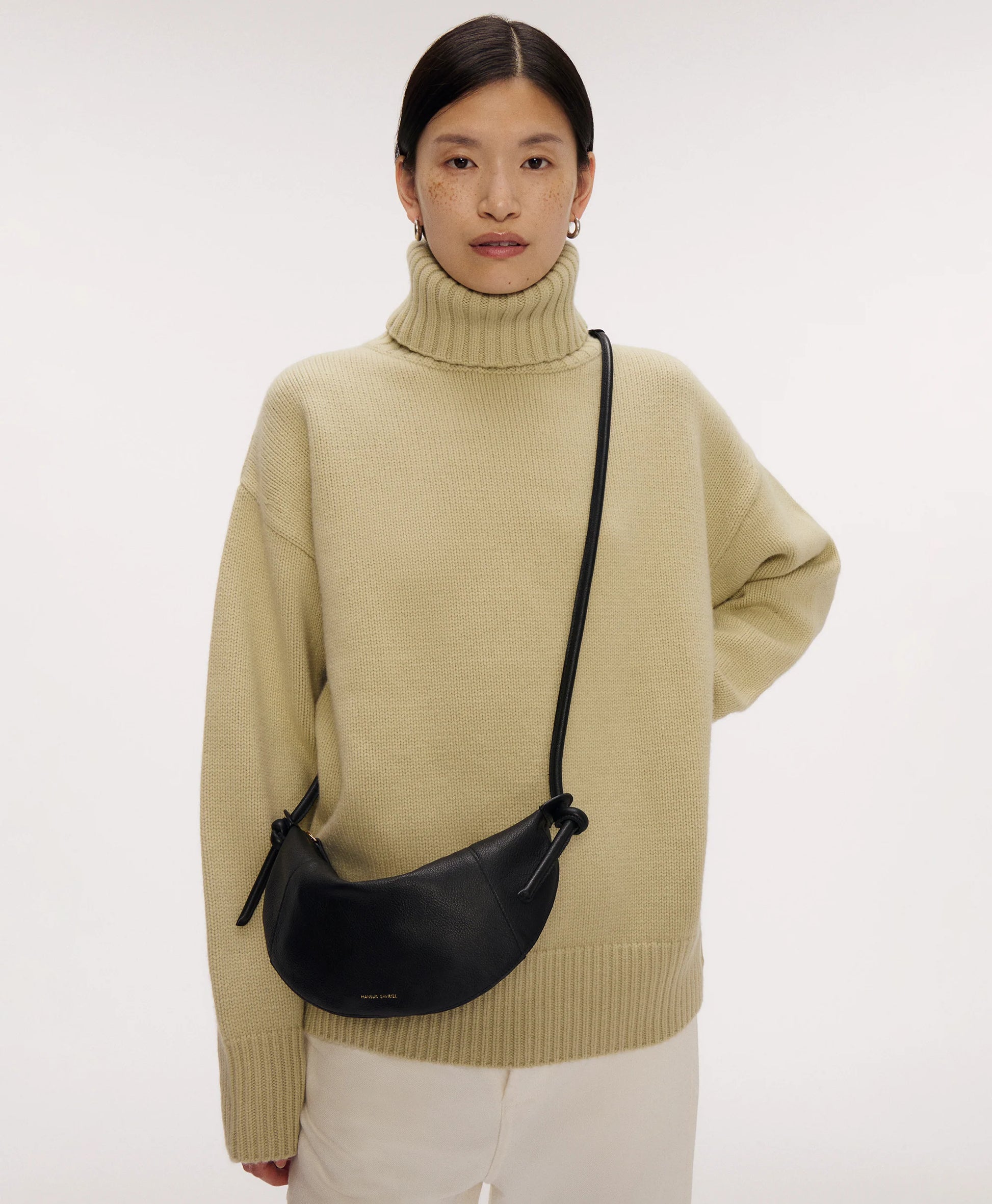 A woman in a beige turtleneck sweater and white pants stands with one hand on her hip, carrying the Mini Fortuna Bag by Mansur Gavriel, crafted from premium calf leather.