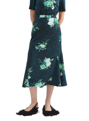 A person wears the Erdem Fluted Abstract Rose Midi Skirt in dark blue with a matching belt, styled with black fringed shoes and hands in pockets.