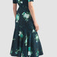 A woman stands with her back to the camera wearing the Erdem Fluted Abstract Rose Midi Skirt paired with black fuzzy flat shoes.