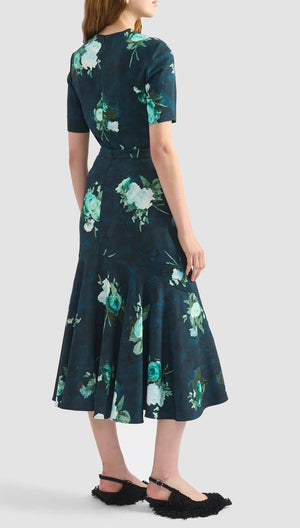 A woman stands with her back to the camera wearing the Erdem Fluted Abstract Rose Midi Skirt paired with black fuzzy flat shoes.