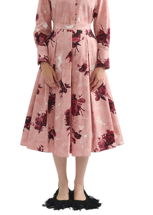 A person wearing Erdem's Pleated Abstract Rose Skirt—a pink, floral midi design—paired with a pink, long-sleeve, collared dress and black fringed shoes, shown from shoulders to feet.