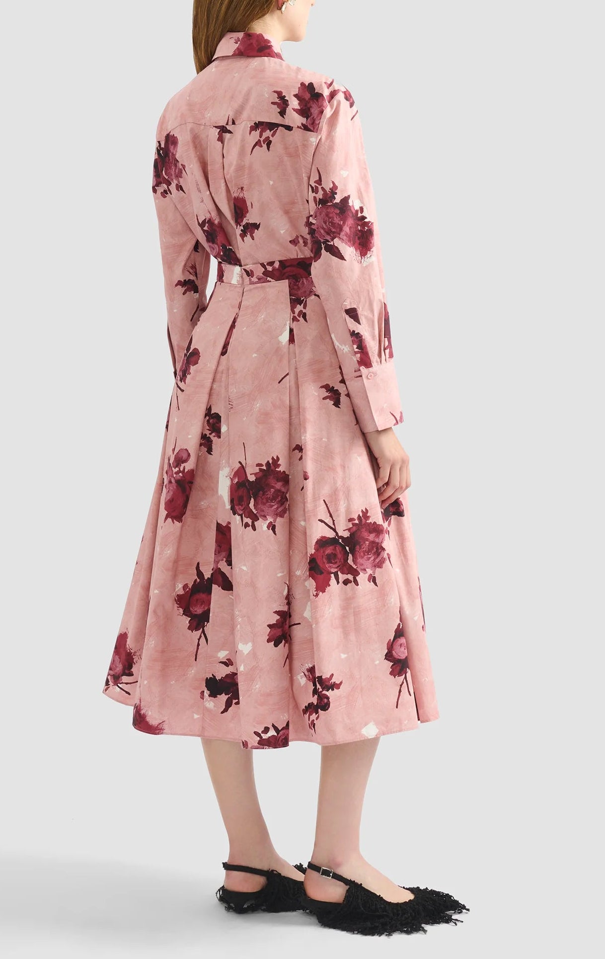 A woman is seen from the back wearing Erdem’s Pleated Abstract Rose Skirt with a coordinating long-sleeve, belted top, and textured black flat shoes.