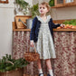 A young girl in a Kidiwi Adeline Smocked Dress and a cardigan stands on a stool in the kitchen. She holds a small basket, surrounded by vegetables and greenery, with the dress's smocked collar adding an extra touch of charm.