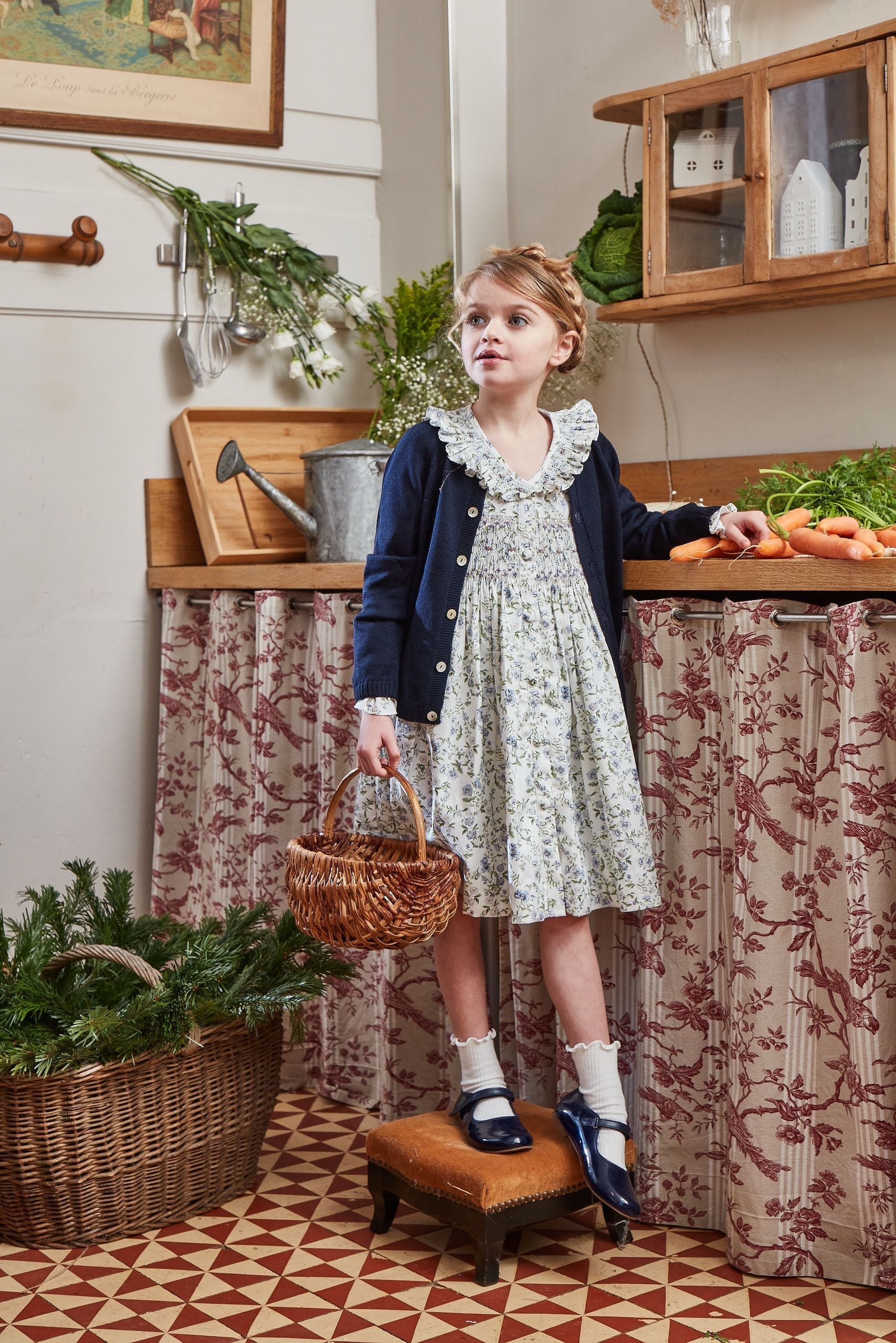 A young girl in a Kidiwi Adeline Smocked Dress and a cardigan stands on a stool in the kitchen. She holds a small basket, surrounded by vegetables and greenery, with the dress's smocked collar adding an extra touch of charm.