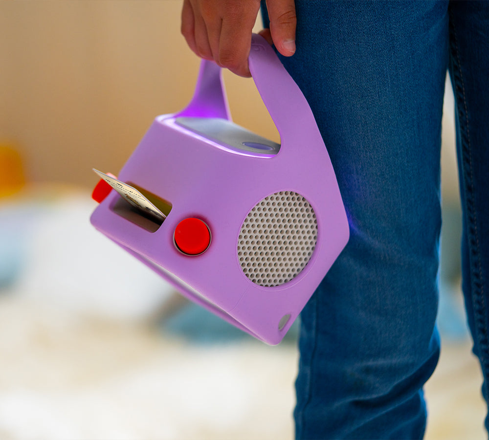 A person holds a Yoto Player, adorned with the Yoto Player Adventure Jacket in purple, featuring a handle, speaker, and card slot, stylishly paired next to blue jeans.