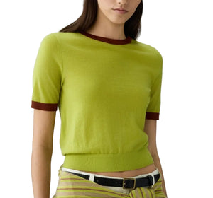 A woman wears the Airy Ringer Tee by Guest in Residence—a vintage-inspired, short-sleeve light green cashmere top with brown trim—paired with striped pants and a black belt.