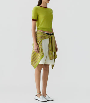 A person wearing the Airy Ringer Tee by Guest in Residence, paired with a white skirt, white shoes, and a striped sweater tied around their waist, stands against a plain background.