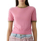 A woman wears the Airy Ringer Tee by Guest in Residence—a vintage-inspired, lightweight pink cashmere short-sleeve sweater with olive green trim—paired with light blue jeans and a pink textured garment tied at her waist.