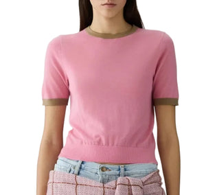 A woman wears the Airy Ringer Tee by Guest in Residence—a vintage-inspired, lightweight pink cashmere short-sleeve sweater with olive green trim—paired with light blue jeans and a pink textured garment tied at her waist.