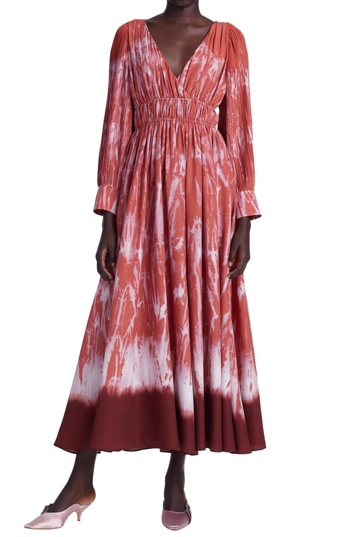 A person wears the Altuzarra Kathleen Dress, a long-sleeved red and white shibori tie-dye dress with an elastic waist, paired with pink high heels, standing gracefully in a studio.