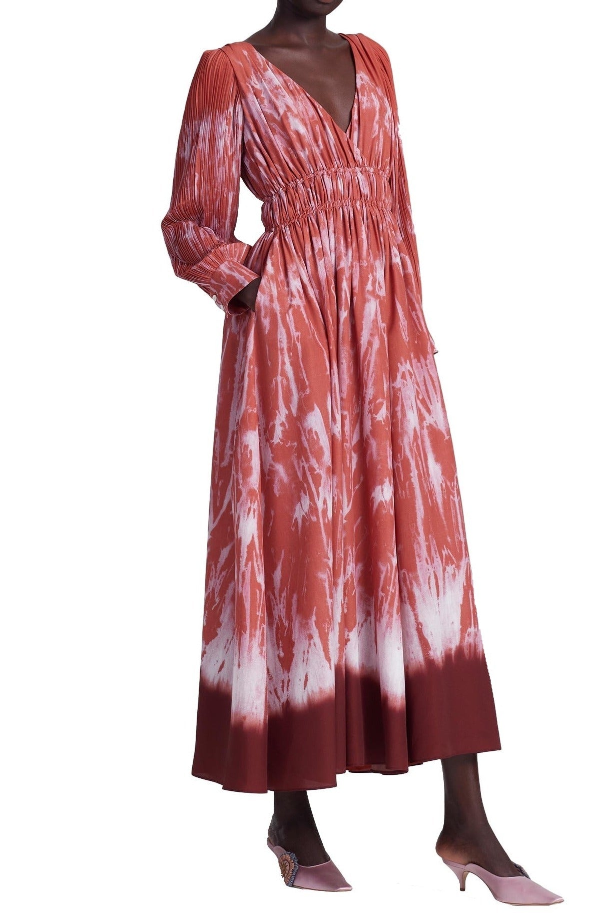 A person is wearing the Altuzarra Kathleen Dress, a long reddish-orange shibori tie-dye dress with an elasticated waist and long sleeves, paired with pink heels.