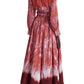 Against a white backdrop, someone models the Altuzarra Kathleen Dress, a long-sleeved garment with red and white shibori patterns and an elasticated waist. The dress pairs beautifully with their short hair and light pink low-heeled shoes.