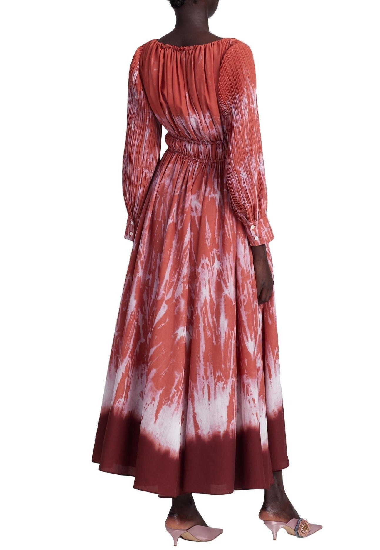 Against a white backdrop, someone models the Altuzarra Kathleen Dress, a long-sleeved garment with red and white shibori patterns and an elasticated waist. The dress pairs beautifully with their short hair and light pink low-heeled shoes.