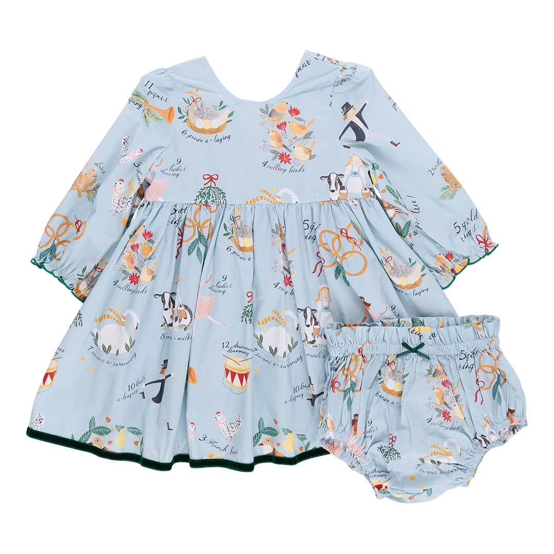 The Pink Chicken Baby Girl Amma Dress showcases a light blue children's dress embellished with festive illustrations such as animals and musical instruments, reminiscent of the "12 Days of Christmas." It includes matching bloomers, making it ideal for your little one's holiday celebrations.