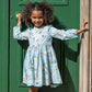 A young girl in the Pink Chicken Girls Amma Dress beams with joy as she holds a green door handle, capturing the festive spirit reminiscent of the 12 Days of Christmas.