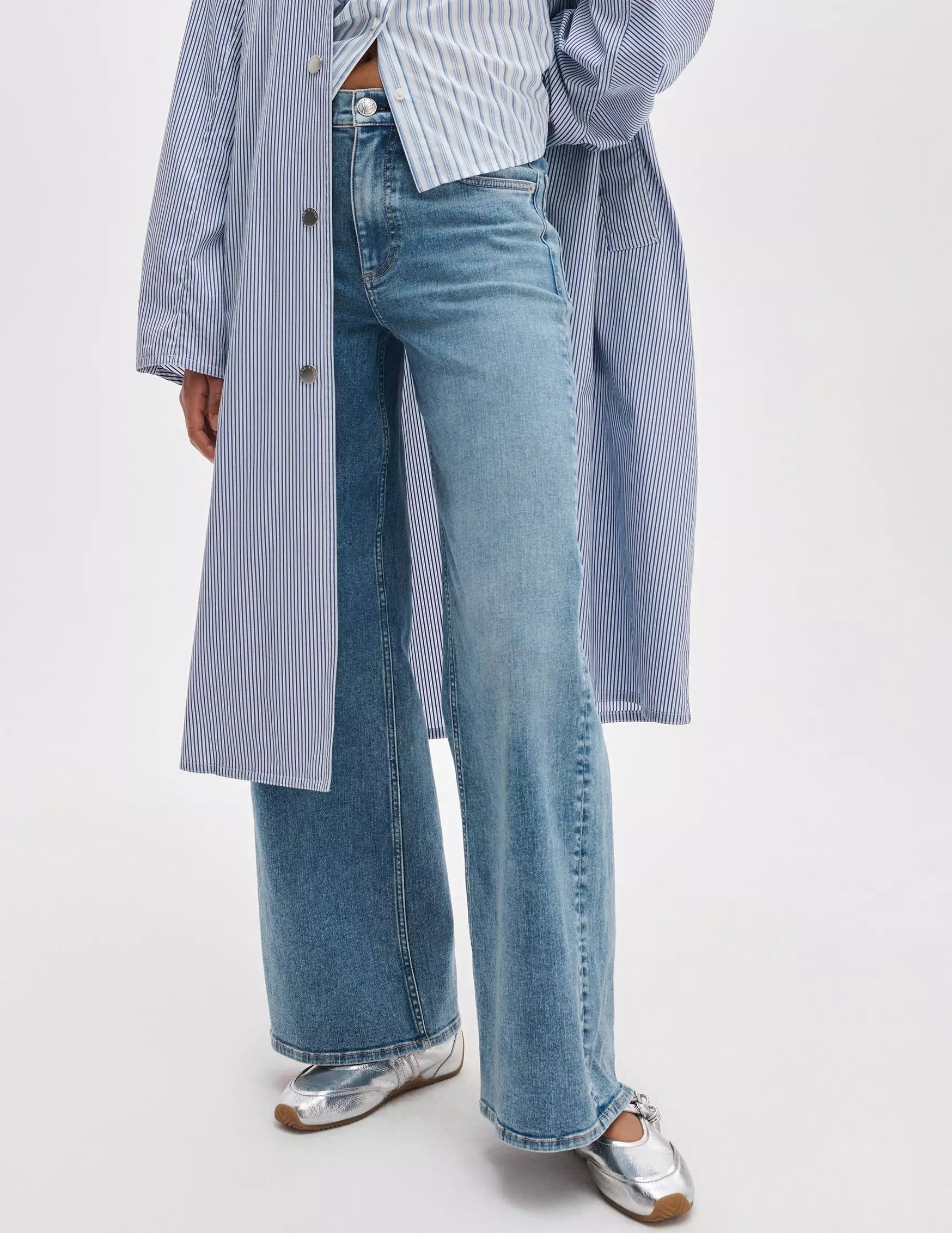 A person wearing Rag & Bone's Flexi Andi Full Wide high-waisted, wide leg jeans, a blue and white striped shirt with a matching long coat, and silver ballet flats stands against a plain background.
