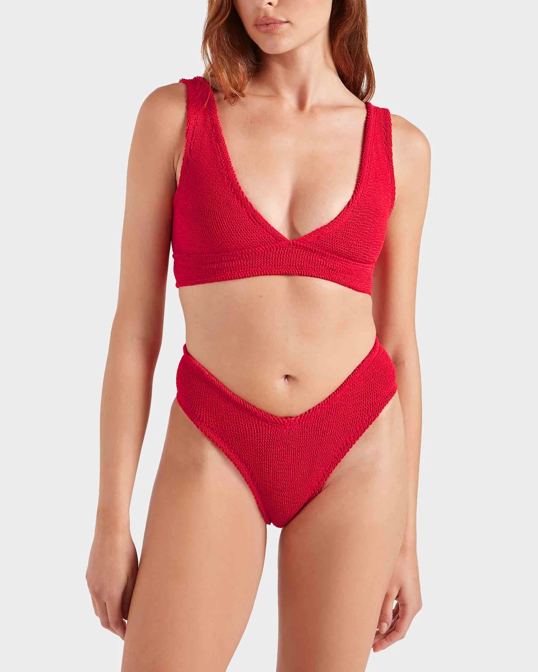 A woman wears the Hunza G Angel Bikini, featuring a textured red V-neck top and high-waisted bottoms, standing against a plain light background.