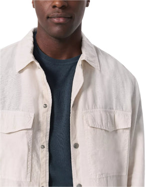 A person wears the Rag & Bone Angus Linen Shirt Jacket—a light-colored, lightweight button-up—over a dark blue textured crew-neck, offering a modern workwear style. Only the subject's shoulders to mouth are visible.