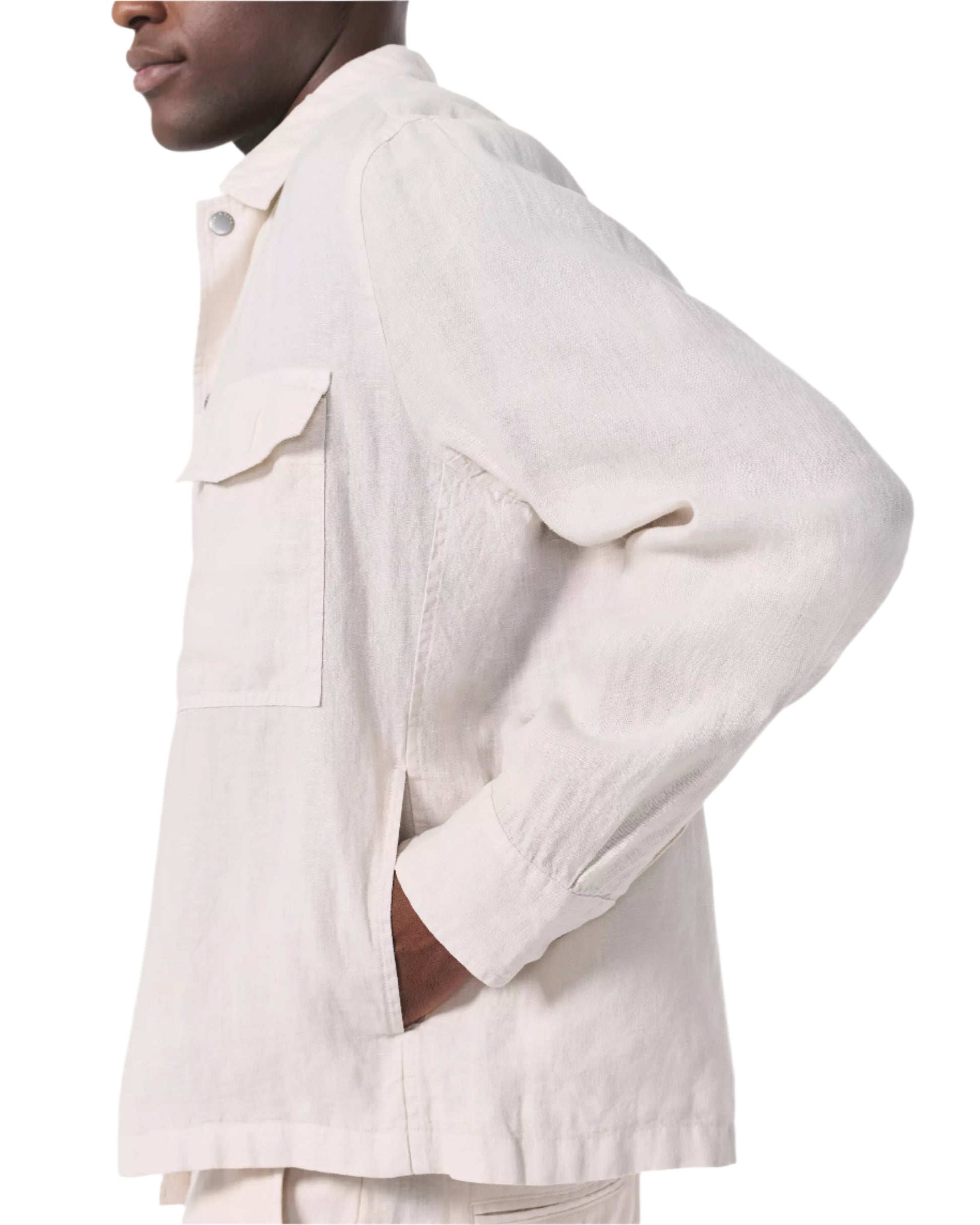 A person stands in side profile with a hand in their pocket, wearing the Rag & Bone Angus Linen Shirt Jacket—an off-white, long-sleeve collared style with a chest pocket made from lightweight linen.