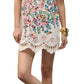 A woman wears the Figue April Dress, a short cream cotton poplin style with colorful floral patterns and scallop embroidery, paired with gold sandals and bracelets, standing with one leg crossed.
