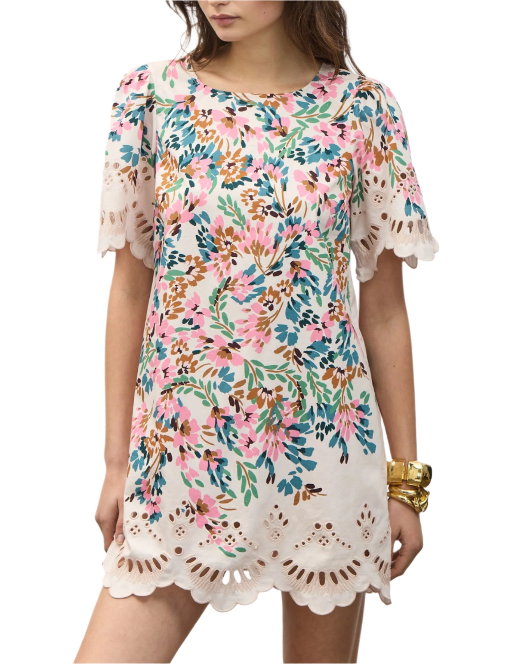A woman wears the Figue April Dress, a short-sleeved cotton poplin floral-print dress with scalloped embroidery at the hem and sleeves in pink, green, and blue, accessorized with gold bracelets.