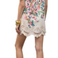 Woman shown from the back wearing the Figue April Dress, a short white Cotton Poplin dress with colorful floral patterns, flutter sleeves, and scallop embroidery at the hem and sleeves. Styled with gold sandals and bracelets.