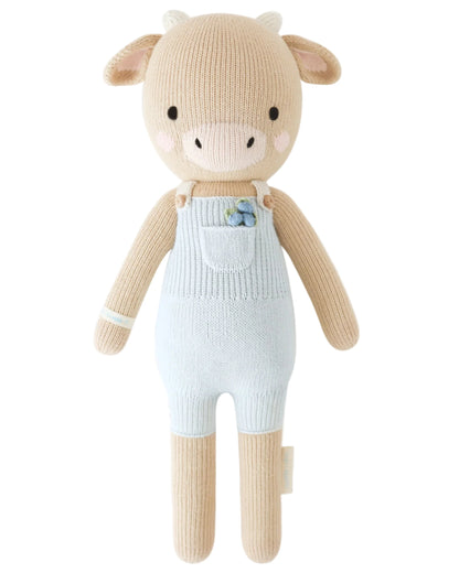 Meet Asher the Cow by Cuddle + Kind, a regular 20" knitted plush in light brown and cream. Wearing pale blue overalls with a flower detail, Asher inspires kindness everywhere. Birthday: May 20th.